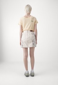 Gul bomulls-T-skjorte med grønn merkevare kombinert med en lys beige denimskjørt med blomstermønster på sidene.