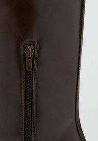 Botte en cuir marron foncé avec une fermeture éclair latérale, une surface texturée, une forme courbée et des détails de couture le long du bord.