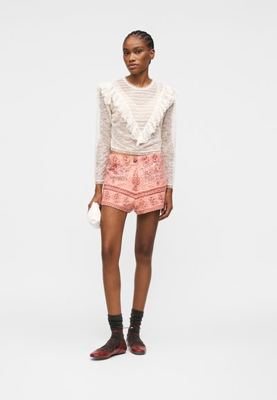 Femme portant un haut à manches longues en dentelle blanche avec des volants, un short à motifs corail, des chaussettes noires, des ballerines rouges, et tenant une petite pochette blanche.