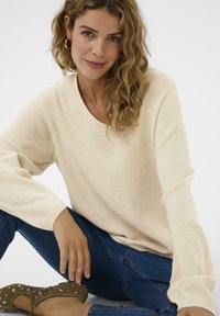 Frau mit welligem Haar, die einen beigen Pullover, blaue Jeans und braune Ballerinas mit Nieten trägt, sitzt mit einem Bein über dem anderen.