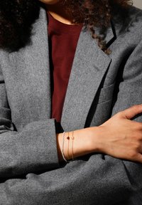 Gold and silver bracelets with a red stone and thin chains, worn on a wrist, against a gray blazer and burgundy shirt background.