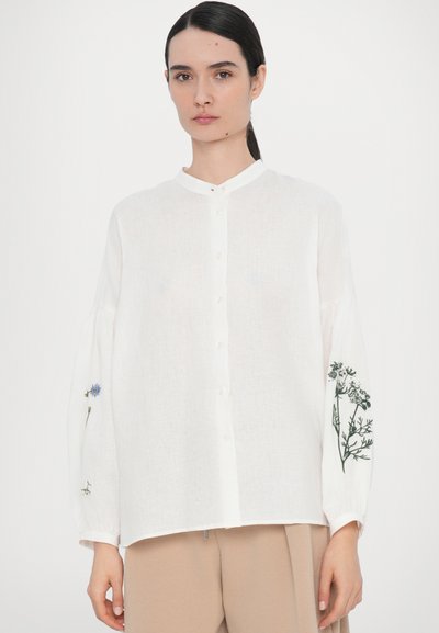 Hvit linne skjorte med ståkrage, knapper foran og broderte blomsterdesign i blått og grønt på ermene.