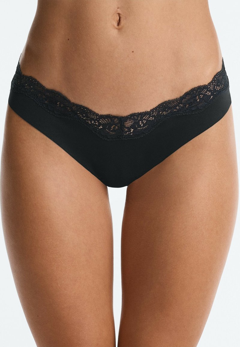 Femme portant un sous-vêtement noir taille basse avec une bordure en dentelle le long de la ceinture, montrée de la taille au haut des cuisses sur un fond clair.