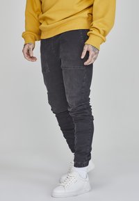 Schwarze, taillierte Jogginghosen mit Seitentaschen, elastischen Bündchen und weicher Textur, kombiniert mit weißen Sneakers und einem gelben Sweatshirt.