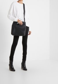 Bolso de hombro negro de cuero con una textura suave, forma rectangular y un asa superior. Llevado por una persona con una blusa blanca y pantalones negros.