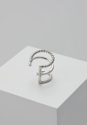 Bague ouverte en argent avec des parties torsadées et lisses, placée sur un cube blanc sur fond gris clair.