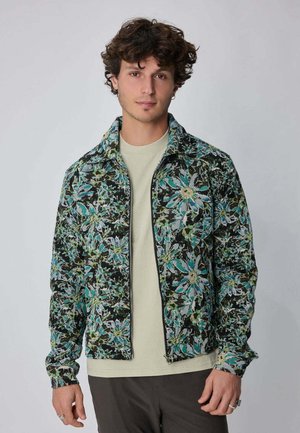 Jeune homme aux cheveux bouclés portant une veste zippée à motifs floraux bleus et verts par-dessus une chemise beige, debout devant un fond gris uni.