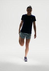 Navy athletic t-shirt, light gray shorts, and purple running shoes. Model standing on one leg, showcasing sporty, functional apparel.
