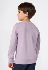 Niño con cabello corto castaño, vistiendo una camiseta de manga larga color púrpura claro y pantalones navy, de pie de perfil contra un fondo blanco.