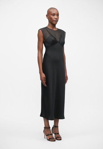 Μαύρο φόρεμα με διαφανές ύφασμα στους ώμους, εφαρμοστό μπούστο και ροή μεσαίας μήκους φούστα. Συνδυασμένο με λεπτά μαύρα ψηλοτάκουνα.