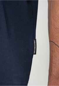 Tamsiai mėlyna medvilninė marškinėliai su juodu etiketu su užrašu "DRAUDŽIAMA", esančiu šone netoli siūlės, su lygiu paviršiumi.