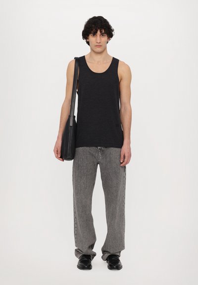 Débardeur noir sans manches, pantalon en denim gris ample et chaussures noires. Le modèle porte également un sac bandoulière noir. Fond blanc uni.