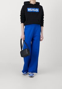 Musta huppari sinisellä "HUGO"-logolla, siniset leveät housut ja pieni musta olkalaukku. Valkoiset ja siniset lenkkarit täydentävät asun.
