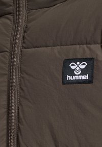 Bruine pufferjack met een ritssluiting, voorzien van een rechthoekig zwart label met een wit logo op de linkerkant van de borst. Gladde textuur.