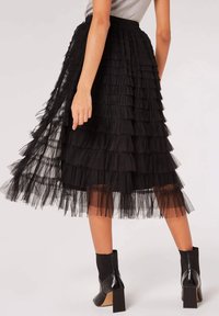 Jupe en tulle noir à volants avec un revêtement transparent, taille élastiquée et ourlet au genou. Associée à des bottines noires.