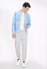Jachetă bomber de un albastru deschis cu un model alb de frunze, purtată cu pantaloni de trening gri și adidași albi. Design simplu și croială lejeră.