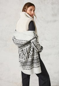 Ljust grå väst med fluffig textur, svart mönstrad sjal hängande över axeln, svarta byxor, står mot en neutral bakgrund.