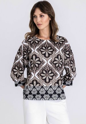 Frau mit braunen, welligen Haaren, die eine locker sitzende Bluse mit schwarz-weißen und beigen floralen sowie geometrischen Mustern trägt, kombiniert mit weißer Hose.