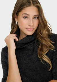Suéter de cuello en cowl de suave y esponjoso tejido negro. Modelo lleva pendientes de declaración plateados, con el cabello castaño ondulado enmarcando el rostro.