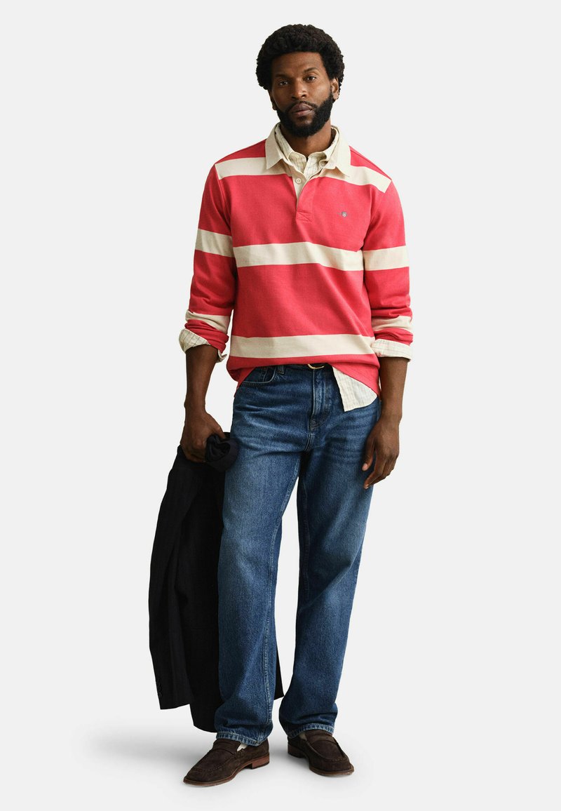 Mann mit Bart, der ein rot-creme gestreiftes Rugby-Shirt, blaue Jeans, braune Schuhe trägt und in der linken Hand eine schwarze Jacke hält.