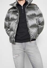 Person iført en skinnende sølv puffjakke over en sort zip-up sweatshirt og lysegrå ripped jeans mod en ensfarvet baggrund.