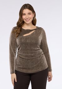 Brązowa, błyszcząca bluzka z długim rękawem, z dekoltem z węzłem i wycięciem w kształcie łezki oraz marszczeniem w talii, zestawiona z ciemnymi spodniami.