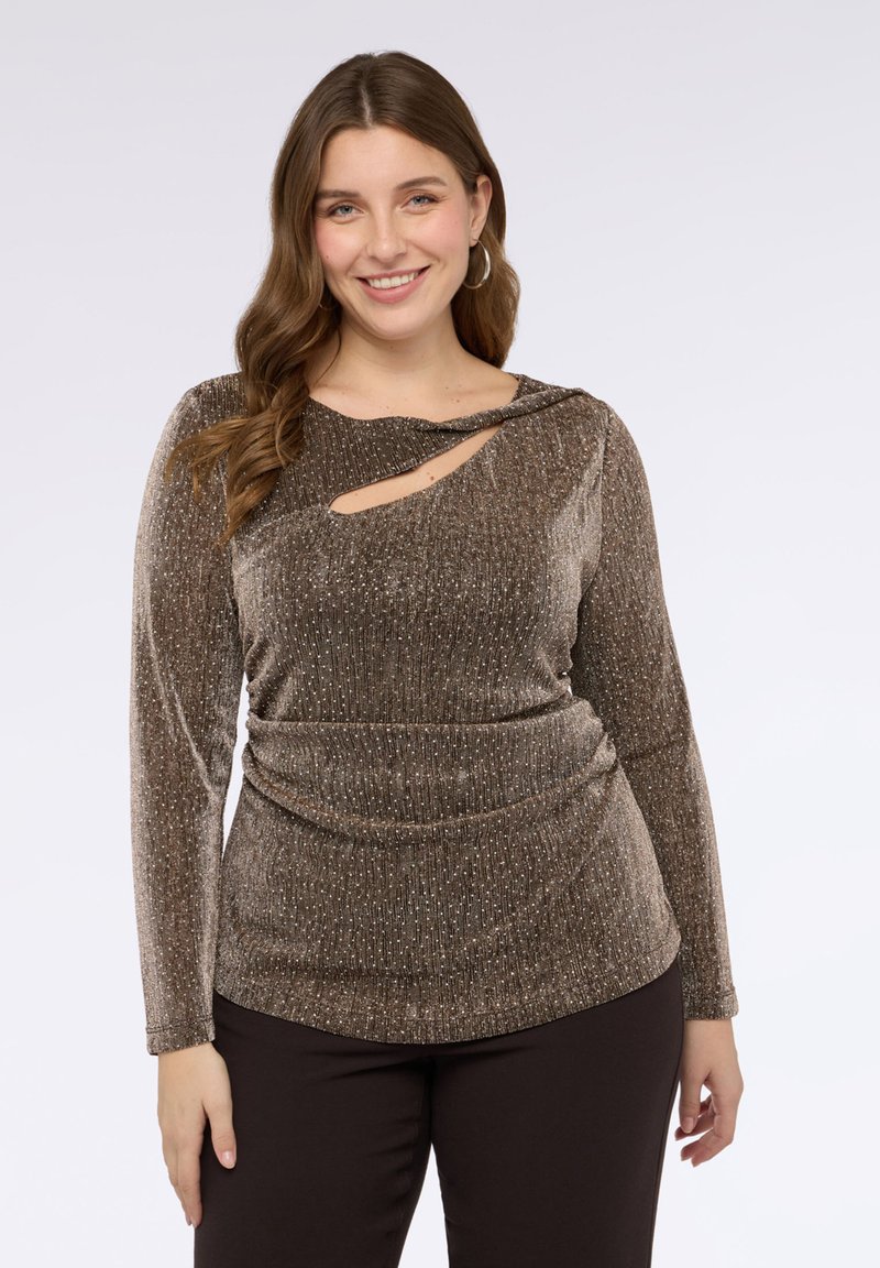 Brązowa, błyszcząca bluzka z długim rękawem, z dekoltem z węzłem i wycięciem w kształcie łezki oraz marszczeniem w talii, zestawiona z ciemnymi spodniami.