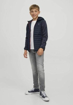 Chaqueta acolchada azul marino con capucha, detalle de cremallera roja, combinada con una camiseta blanca, jeans gris claro y zapatillas azul marino con suelas blancas.