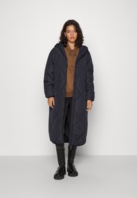 Manteau matelassé long et bleu marine foncé avec une capuche, présentant un motif diagonal et une fermeture éclair sur le devant. Porté sur un sweat à capuche marron et un pantalon noir.