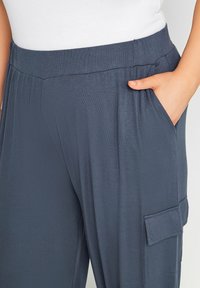 Person som bär mörkblå byxor med elastisk midja och sid- samt cargo-fickor, hand vilande i sidofickan, iförd en vit topp.