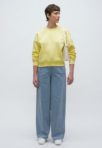 Κίτρινο φαρδύ φούτερ, ανοιχτό γαλάζιο τζιν με φαρδιά πατζάκια, λευκά αθλητικά παπούτσια και μικρή κρεμ τσάντα χειρός. Απλός, καθημερινός σχεδιασμός με άνετη εφαρμογή.