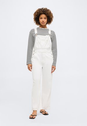 Mladá žena s kučeravými vlasmi, ktorá má na sebe čierno-biele pruhované tričko s dlhým rukávom pod bielymi rifľovými náprsenkami a čierne sandále s otvorenou špicou.