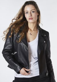 Giubbotto da moto in pelle nera con accenti in metallo argentato, design aderente e cerniere. Indossato sopra una maglietta bianca a scollo a V.