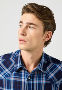 Camicia a scacchi blu con un motivo di quadrati blu scuro, blu chiaro e arancione. Collo button-up e dettaglio della spalla visibile.