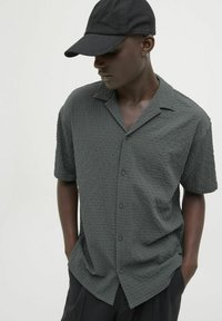 Camisa de manga corta y cuello en tejido de color gris oscuro con botones en la parte delantera. Combinada con una gorra negra y pantalones negros sueltos.