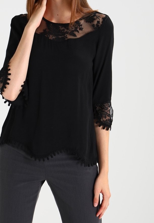 Femme portant un chemisier noir avec de la dentelle florale transparente au niveau du col et des manches, associé à un pantalon gris foncé, debout devant un fond uni.