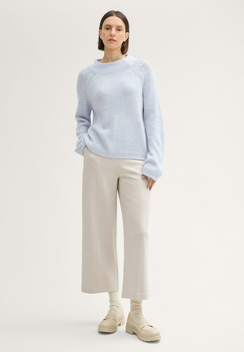 Jasnoniebieski sweter dzianinowy z prążkowanymi mankietami, w połączeniu z beżowymi spodniami o szerokich nogawkach i kremowymi butami na platformie. Całość jest prosta i nowoczesna.