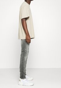 Camiseta oversized beige, jeans ajustados grises y zapatillas blancas. La vista lateral resalta el ajuste relajado de la camiseta y el denim texturizado.