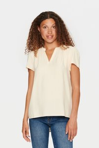 Blusa corta a maniche corte color crema chiaro con scollo a V, tessuto tessurizzato, dettaglio con ruffle sul collo, abbinata a jeans in denim blu.