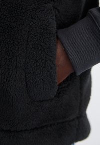 Veste en polaire noire avec une surface texturée et fluffy. Dispose d'une poche kangourou et de poignets côtelés dans une teinte plus sombre, ajoutant à la chaleur et au confort.