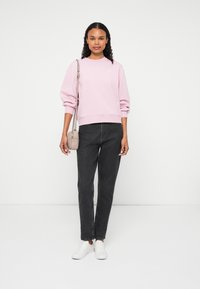Sweatshirt roz deschis cu manșete cu nervuri, asortat cu blugi negri și adidași albi. Modelul ține o geantă mică bej tip crossbody.