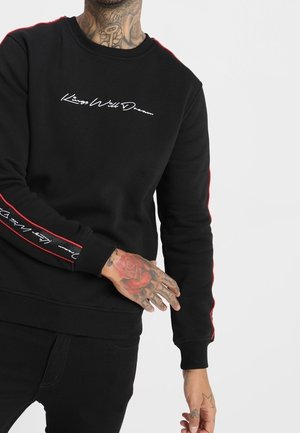 Person iført sort sweatshirt med hvid skrift "Kiss Well Dream" på bryst og ærme, der viser tatoveret hånd og nakke.