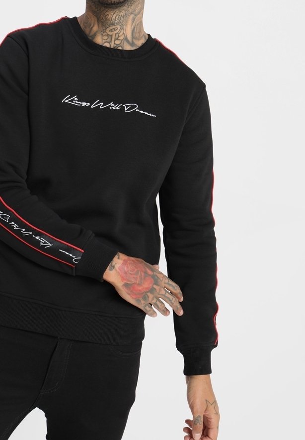Personne portant un sweat-shirt noir avec le texte en écriture blanche « Kiss Well Dream » sur la poitrine et la manche, montrant une main et un cou tatoués.