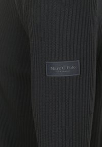 Schwarzer gerippter Pullover mit strukturiertem Strick, versehen mit einem kleinen rechteckigen Etikett mit der Aufschrift "Marc O'Polo" am Ärmel. Klassisches Design.