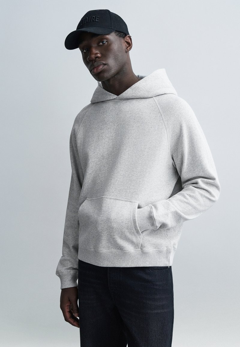 Šedá bavlněná mikina s kapucí, přední kapsou a širokou kapucí. Model ji nosí s tmavými džínami a černou čepicí s vyraženým textem.