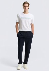 T-shirt blanc en coton avec texte bleu, associé à un pantalon de survêtement bleu marine avec cordon de serrage, tissu texturé, et des baskets blanches.