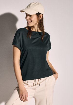 Dunkelgrünes T-Shirt mit kurzen Ärmeln in lässiger Passform und strukturiertem Schulterdetail, kombiniert mit hellbeigen Hosen und einer hellen Kappe.