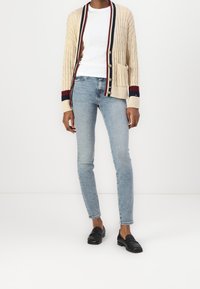 Cardigan en tricot beige avec un motif texturé, rayures rouges et marines sur les poignets, et poches latérales ; associé à un jean bleu clair et des mocassins noirs.
