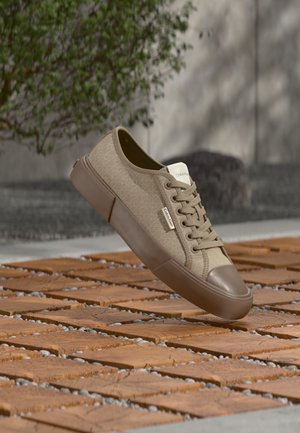 Beige Calvin Klein sneaker med gummisål og snørebånd, svævende over et udendørs trægulv med grøn plante i baggrunden.