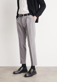 Grå plisserade byxor med skräddarsytt snitt, kombinerade med ett svart läderbälte och svarta klädkor, över en vit skjorta.